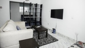 A modern living room with a white sofa, a black coffee table, a black bookshelf, and a flat-screen TV mounted on the wall. - Capital & County Apartment Ikate (Lekki)