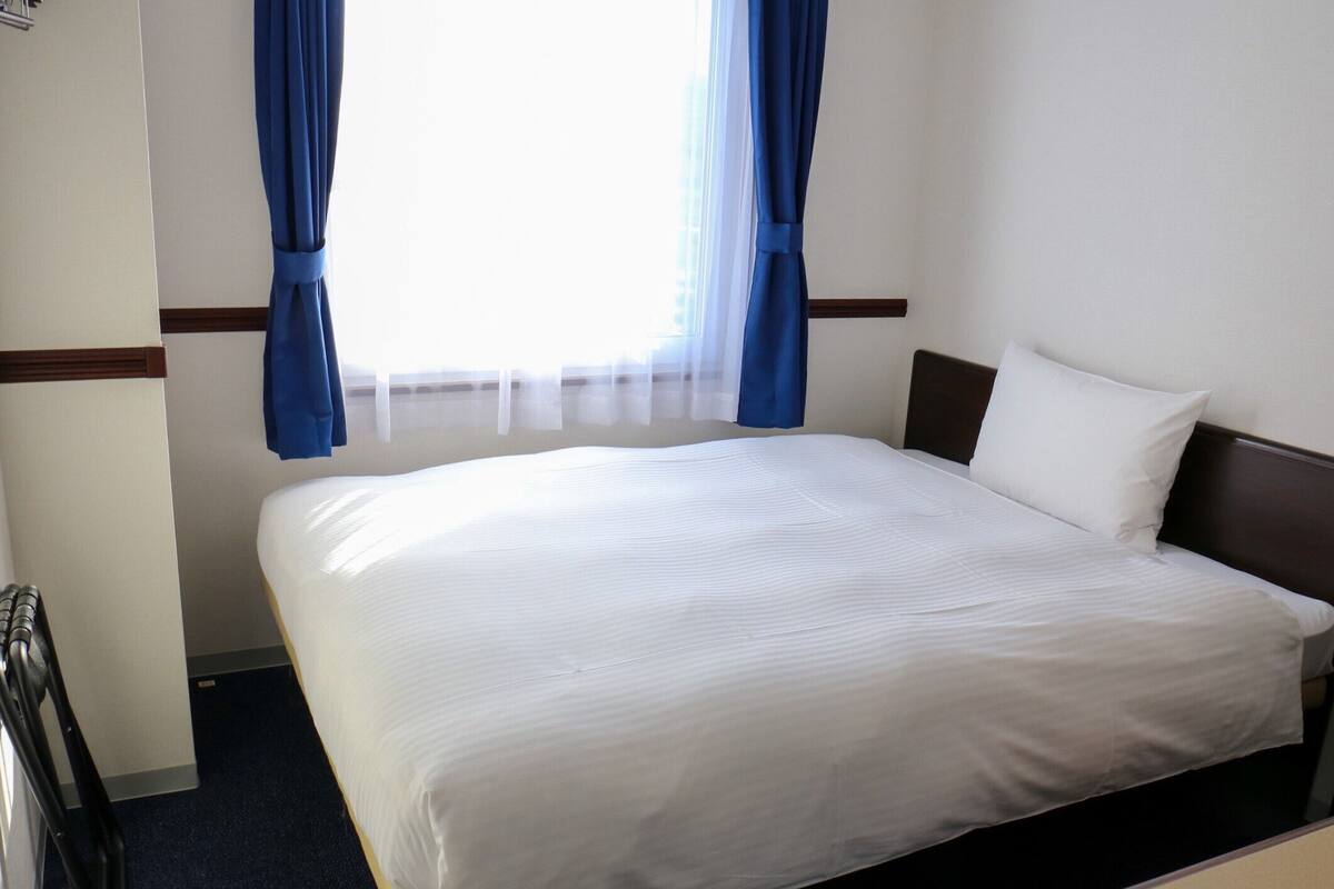 Habitación de hotel con una cama grande, cabecera de madera, mesita de noche y ventana con cortinas azules.