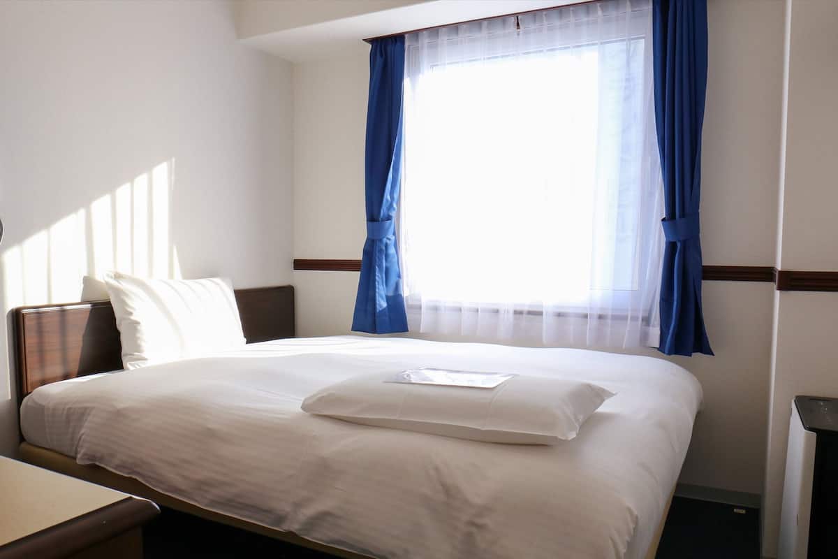 Una habitación de hotel con una cama bien hecha, una mesita de noche y una ventana con cortinas azules.