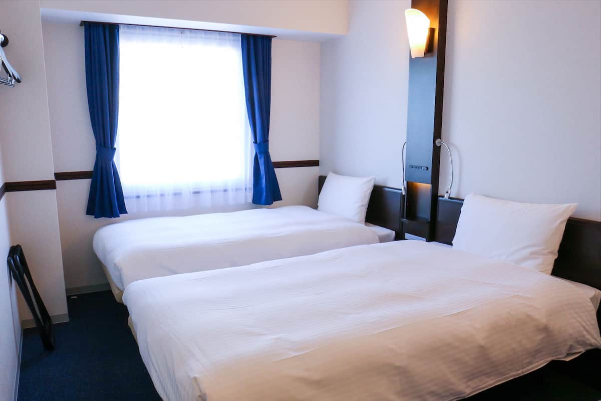 Una habitación de hotel con dos camas, una ventana con cortinas azules, una mesita de noche y una lámpara fijada a la pared.