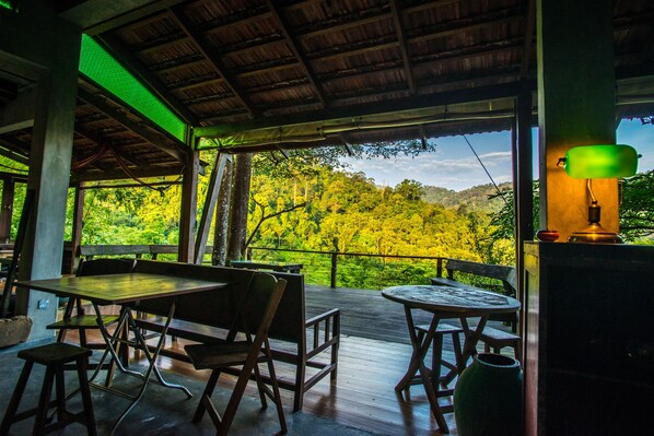 An outdoor dining area with wooden tables and chairs, a covered roof, and a view of a forested hillside. - Spyder Hill (Seremban)
