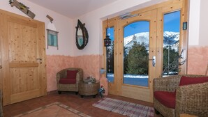 A cozy room with a large wooden door, wicker furniture, a small table, and a view of snow-capped mountains through the door. - Almhaus Blümel (Reichenau)