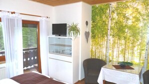 A bedroom with a bed, a TV, a small table with a plant, and a view of trees through a window. - Almhaus Blümel (Reichenau)