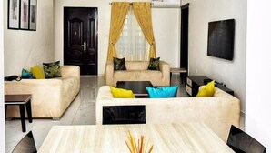 A modern living room with two sofas, a coffee table, and a dining table. There is a door with a dark frame and yellow curtains, and an air conditioning unit mounted on the wall. - Madiba Court (Lekki)