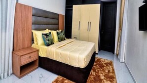 A bedroom with a bed, nightstand, wardrobe, and a brown rug. - Madiba Court (Lekki)