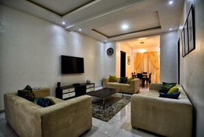 A living room with a flat-screen TV, two sofas, a coffee table, and a dining area with a table and chairs. - Madiba Court (Lekki)