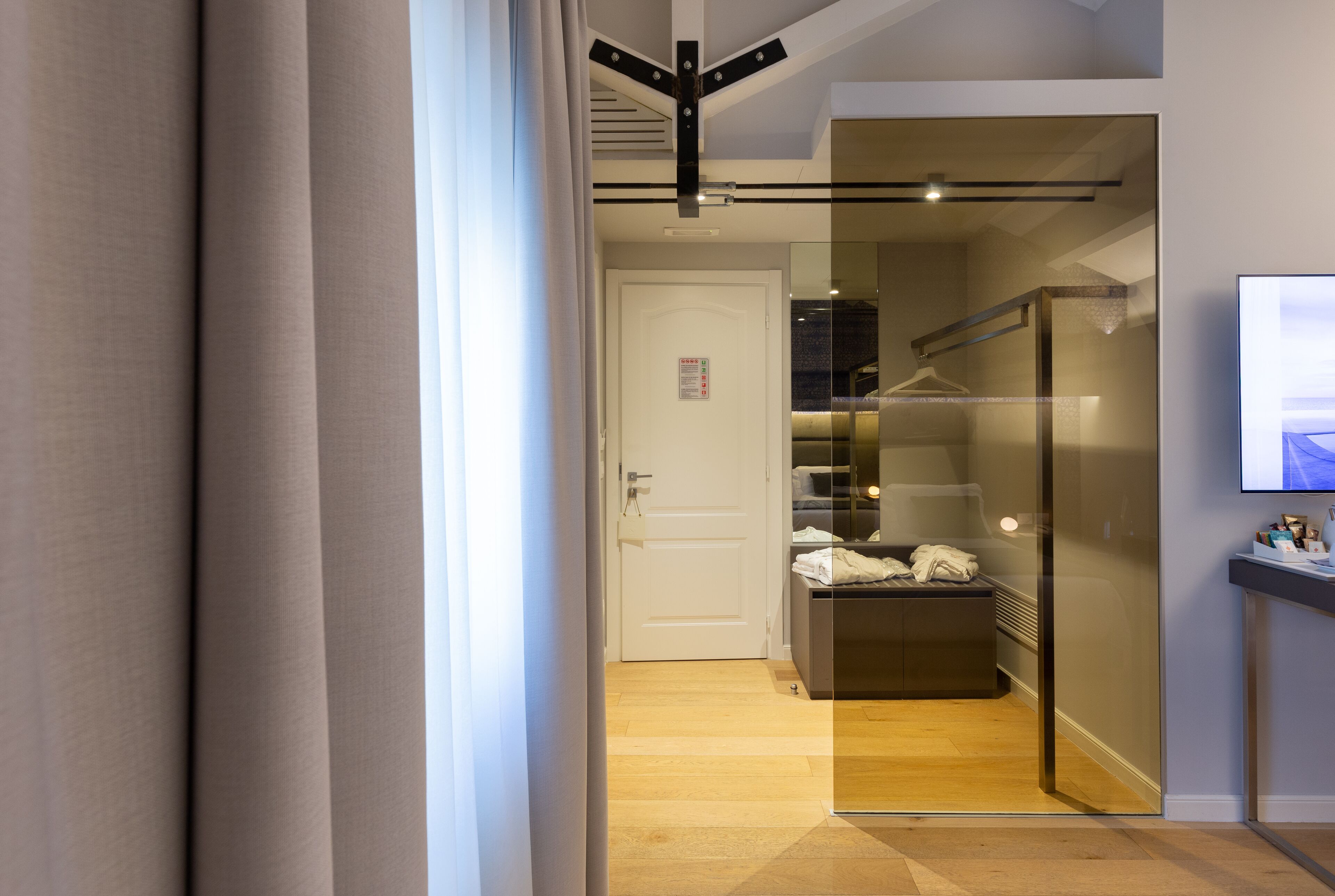 A modern hotel room with a glass partition, a wooden floor, and a visible closet with hanging clothes.