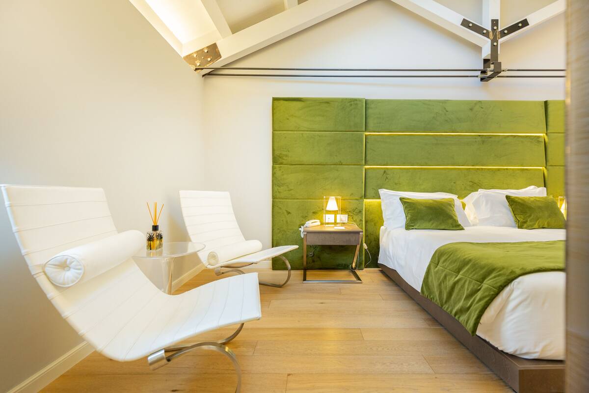 A modern hotel room with a green headboard, a white bed, a white chair, and a bedside table with a lamp.