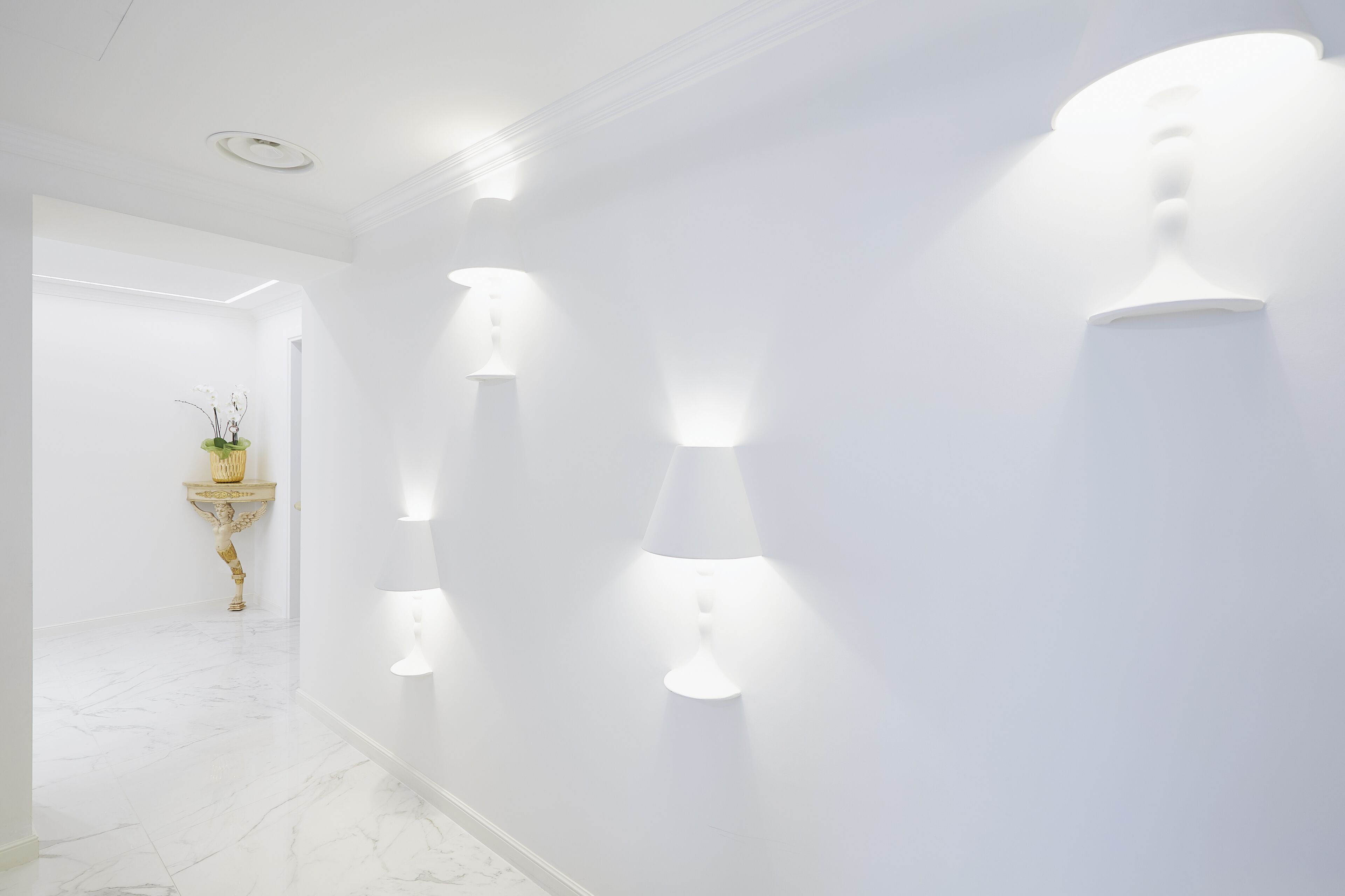 Un corridoio con pareti bianche e pavimento in marmo, illuminato da luci a parete e decorato con una pianta.