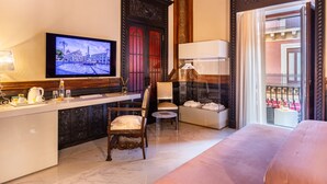 A hotel room with a TV, a dining area with a table and chairs, a sofa, and a balcony with a railing.
