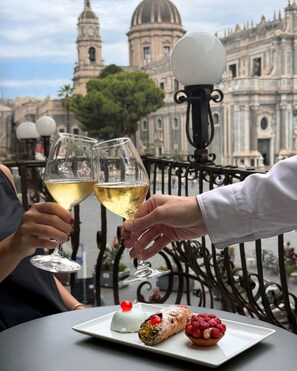 Una persona sosteniendo dos copas de vino, un plato de postre con un pastel y frutos rojos, y un edificio histórico con una cúpula al fondo.