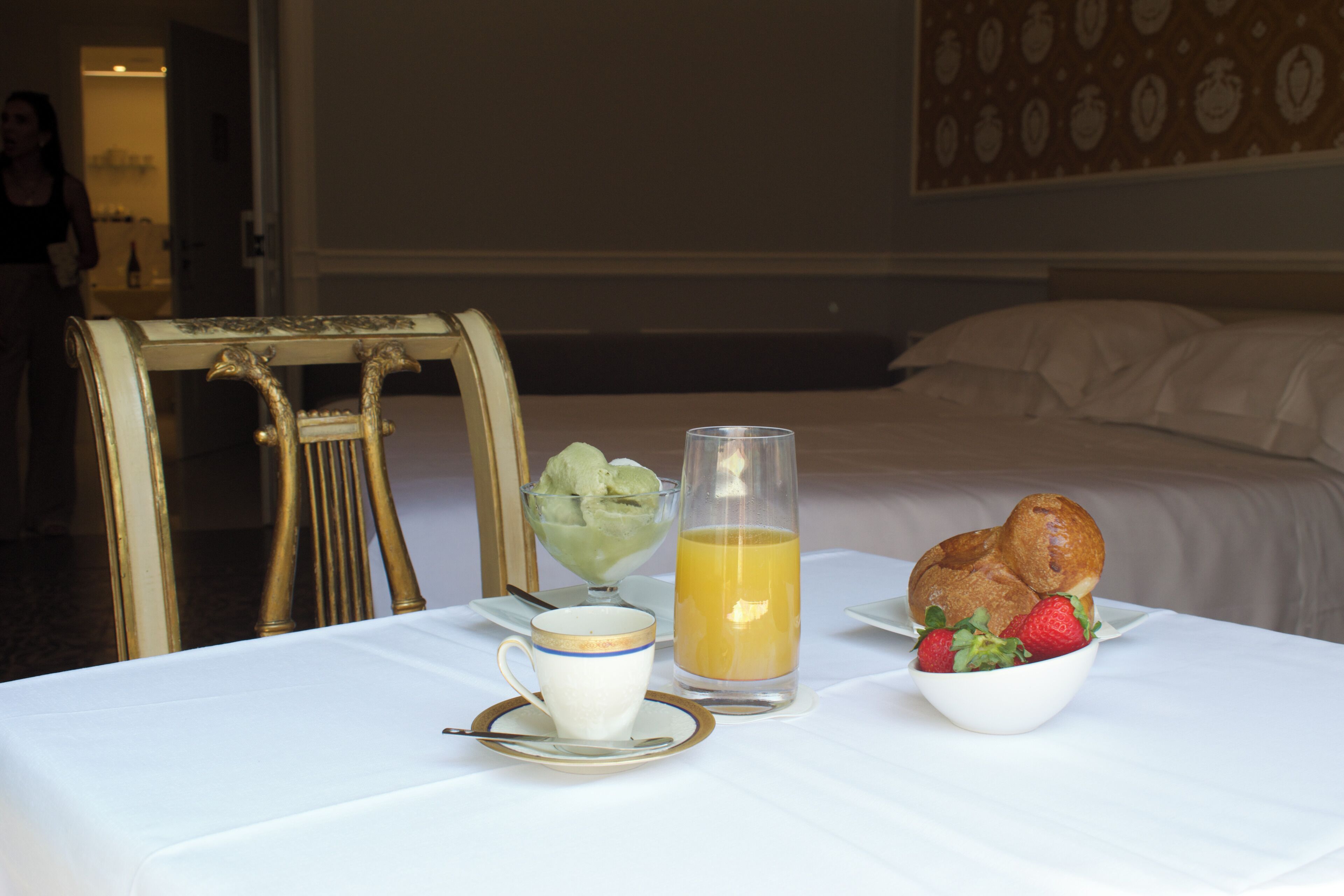 A breakfast setup with a cup, a glass of orange juice, a bowl of strawberries, and a pastry on a white tablecloth.