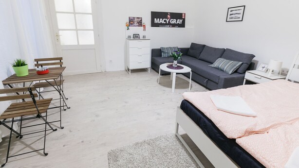 A modern, well-lit room with a grey sofa, a small white table, a wooden bench, and a bed with a pink blanket. - Classic Ernesto near Opera (Budapest)