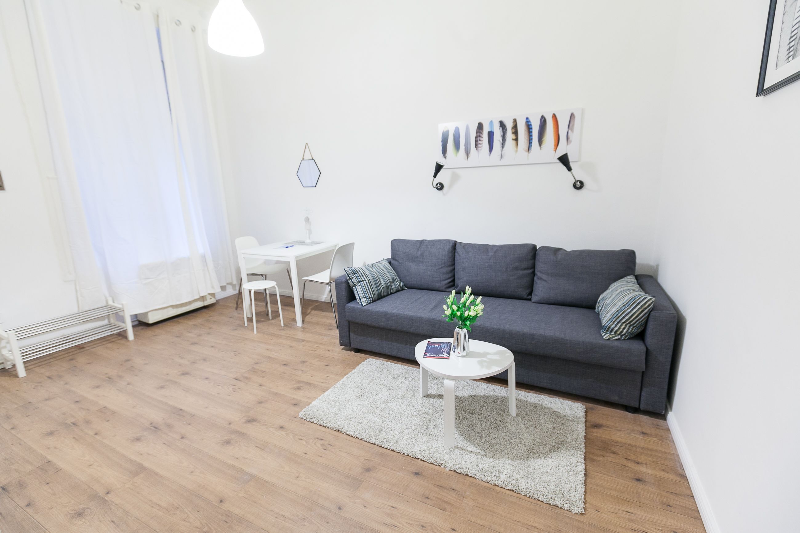 A modern living room with a grey sofa, a white coffee table, and a white chair. There is a white radiator, a white curtain, and a wall-mounted shelf with decorative items.