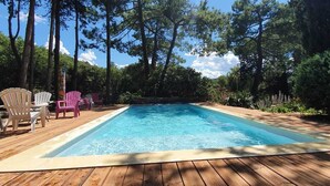 Una terraza de madera con piscina, rodeada de árboles y mobiliario exterior.