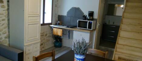 A compact kitchen with a microwave, a kettle, and a small refrigerator. There is a dining table with chairs and a vase with dried flowers.