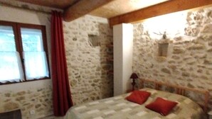 Un dormitorio con pared de piedra, una cama con colcha, cabecero de madera, mesita de noche, ventana con cortinas y una pequeña mesa.