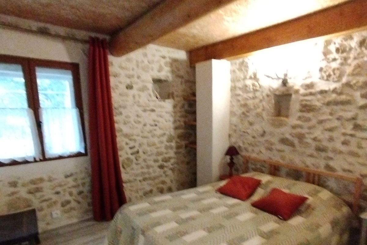 A bedroom with a stone wall, a bed with a quilt, a wooden headboard, a nightstand, a window with curtains, and a small table.