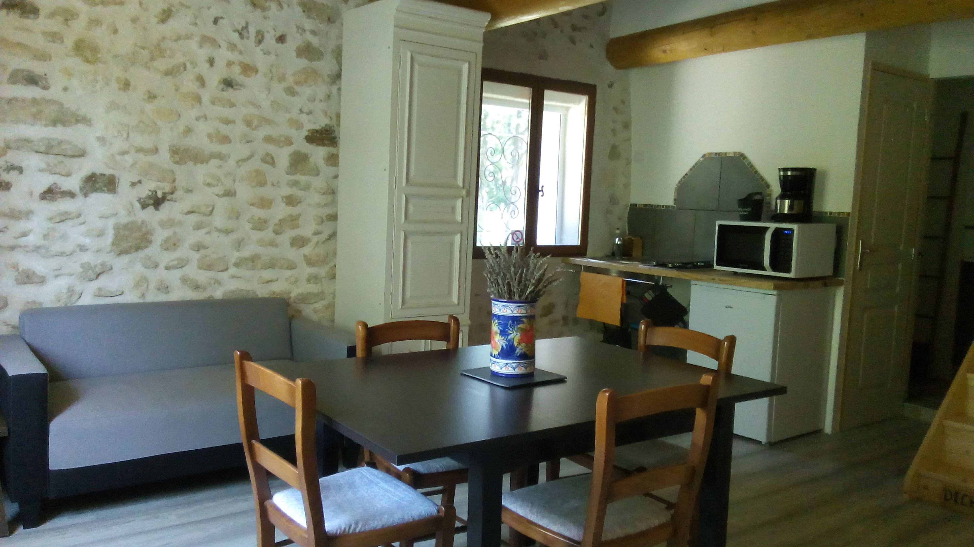 A dining area with a table, chairs, and a vase of dried plants. There is a kitchen area with a microwave and a sink.