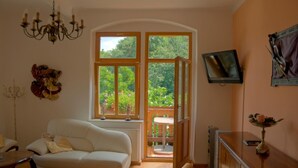 A room with a white sofa, a chandelier, a TV mounted on the wall, and a large window with a view of greenery. - Domizil Adler (Dresden)