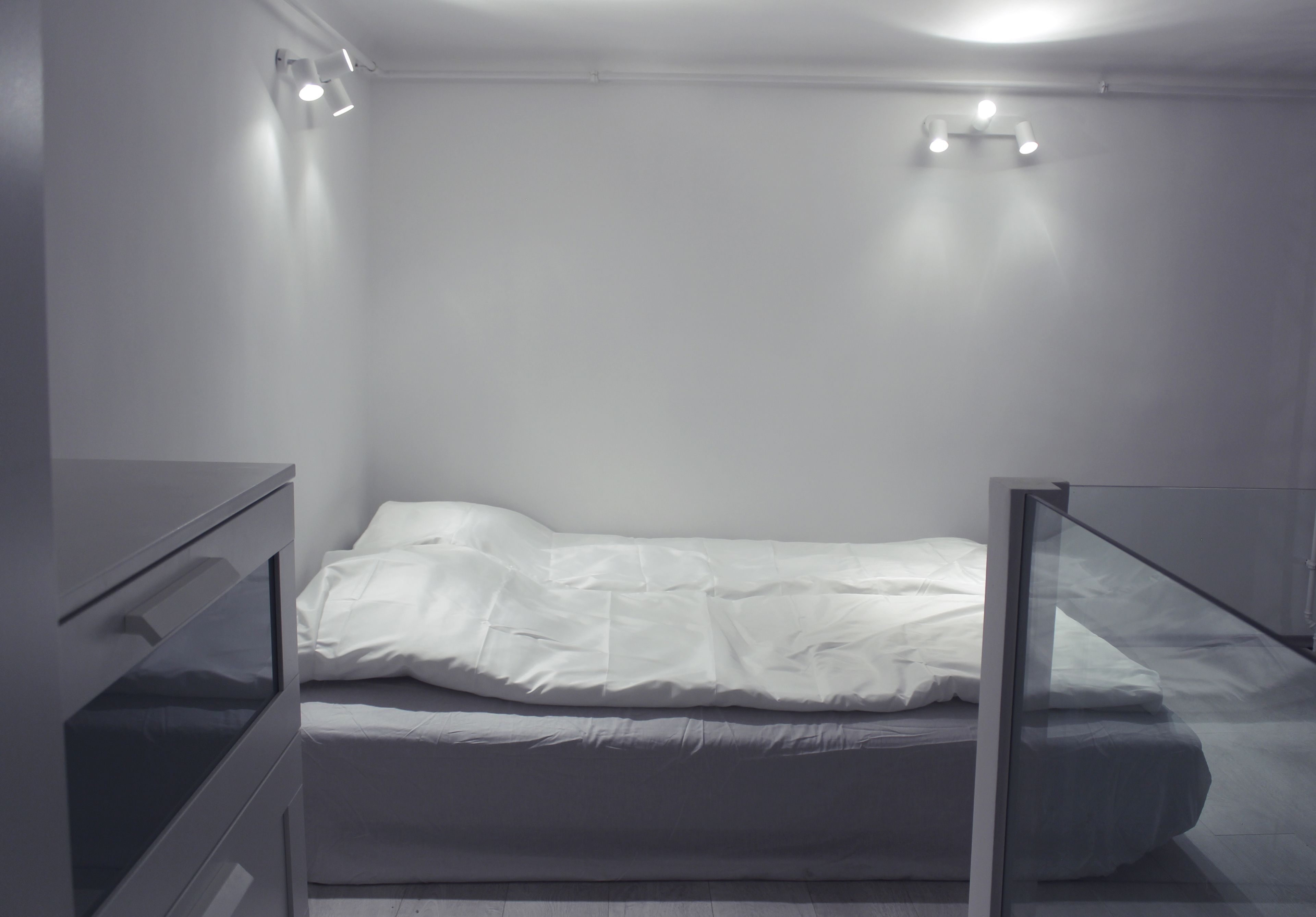 Un llit individual amb roba de llit blanca a una habitació amb parets blanques i una separació de vidre.