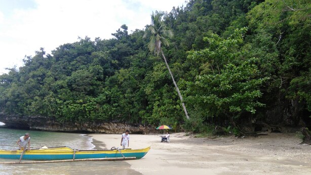 A beach with a boat, a person standing on the boat, and another person sitting under an umbrella on the shore. - Western Highway Lodge (Marabut)