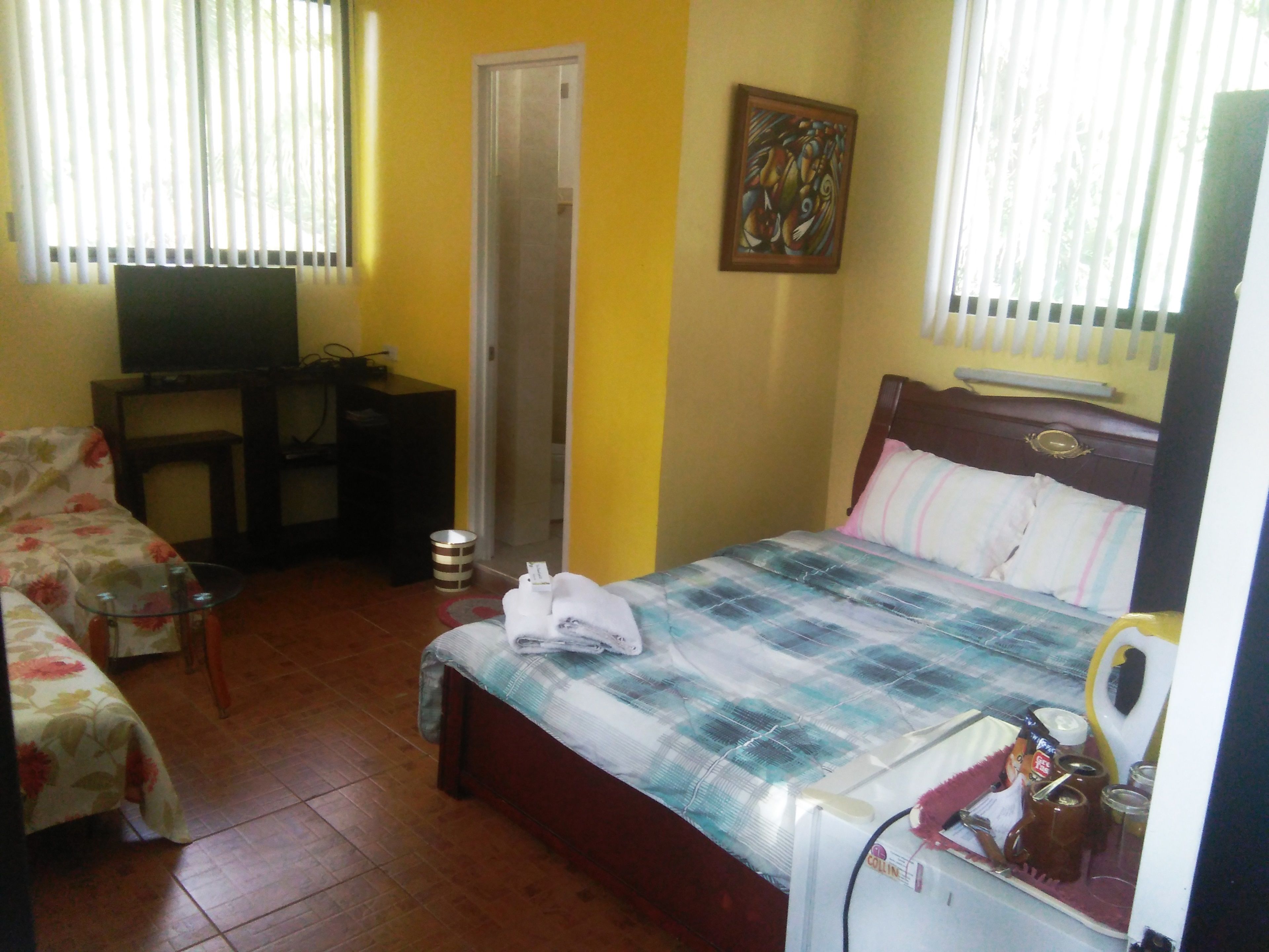 A bedroom with a bed, a desk, a chair, a television, and a window with blinds.