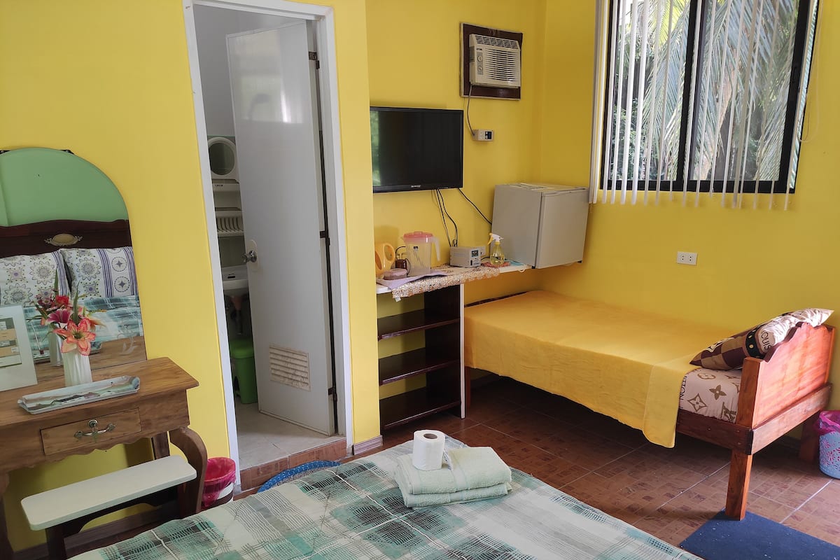 Una habitación pequeña con paredes amarillas, que contiene una cama, un escritorio, una silla, un televisor y una ventana con persianas.