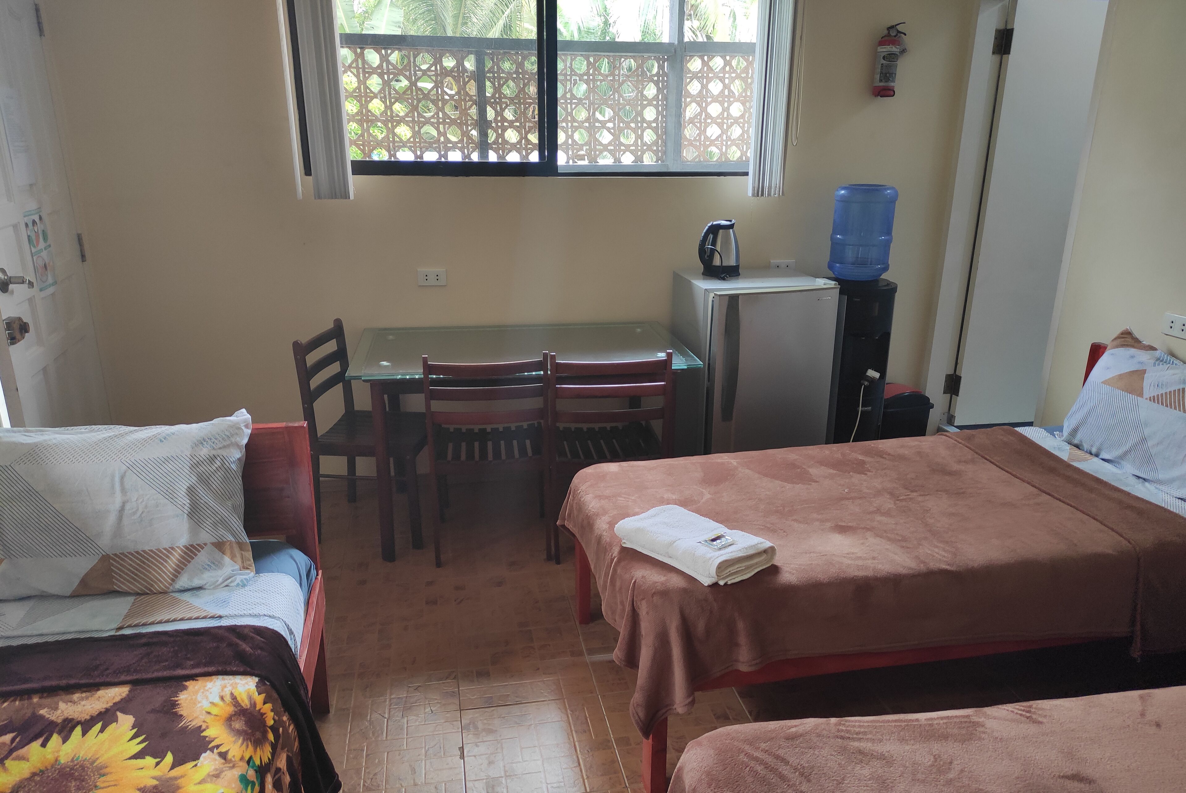 Habitación con dos camas, una mesa pequeña con sillas, un dispensador de agua y una ventana que da a zonas verdes.