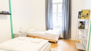 Una habitación de hotel con dos camas, una mesita de noche, un estante con libros y una ventana con cortinas.