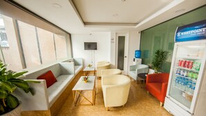 A modern hotel lobby with a seating area, a television, a refrigerator with beverages, and a large window.