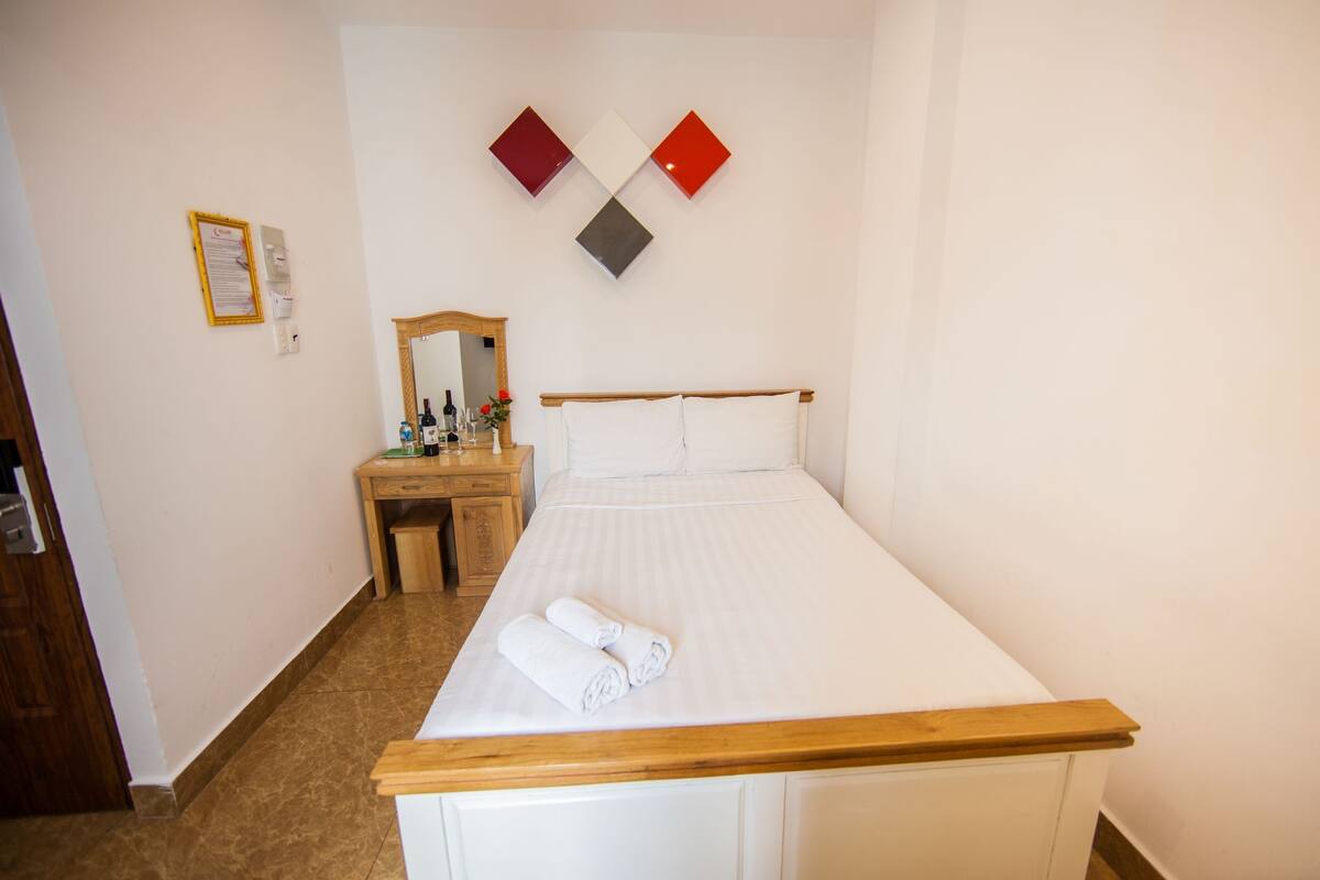 Una habitación de hotel con una cama, cabecera de madera, mesita de noche con espejo y un escritorio pequeño con artículos de aseo.