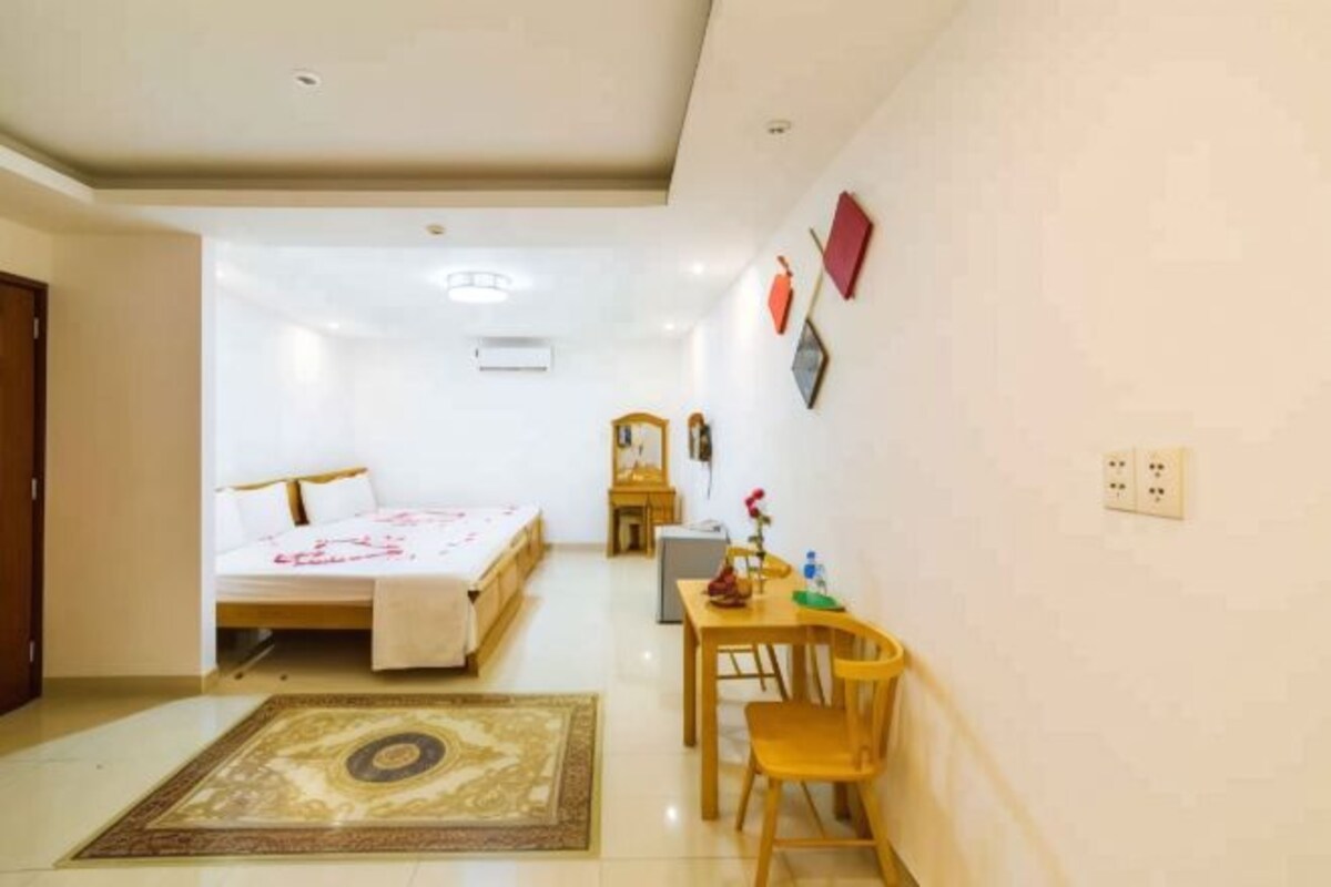 Une chambre d’hôtel comprenant un lit, un bureau avec une chaise, une petite table avec un vase et une étagère fixée au mur.