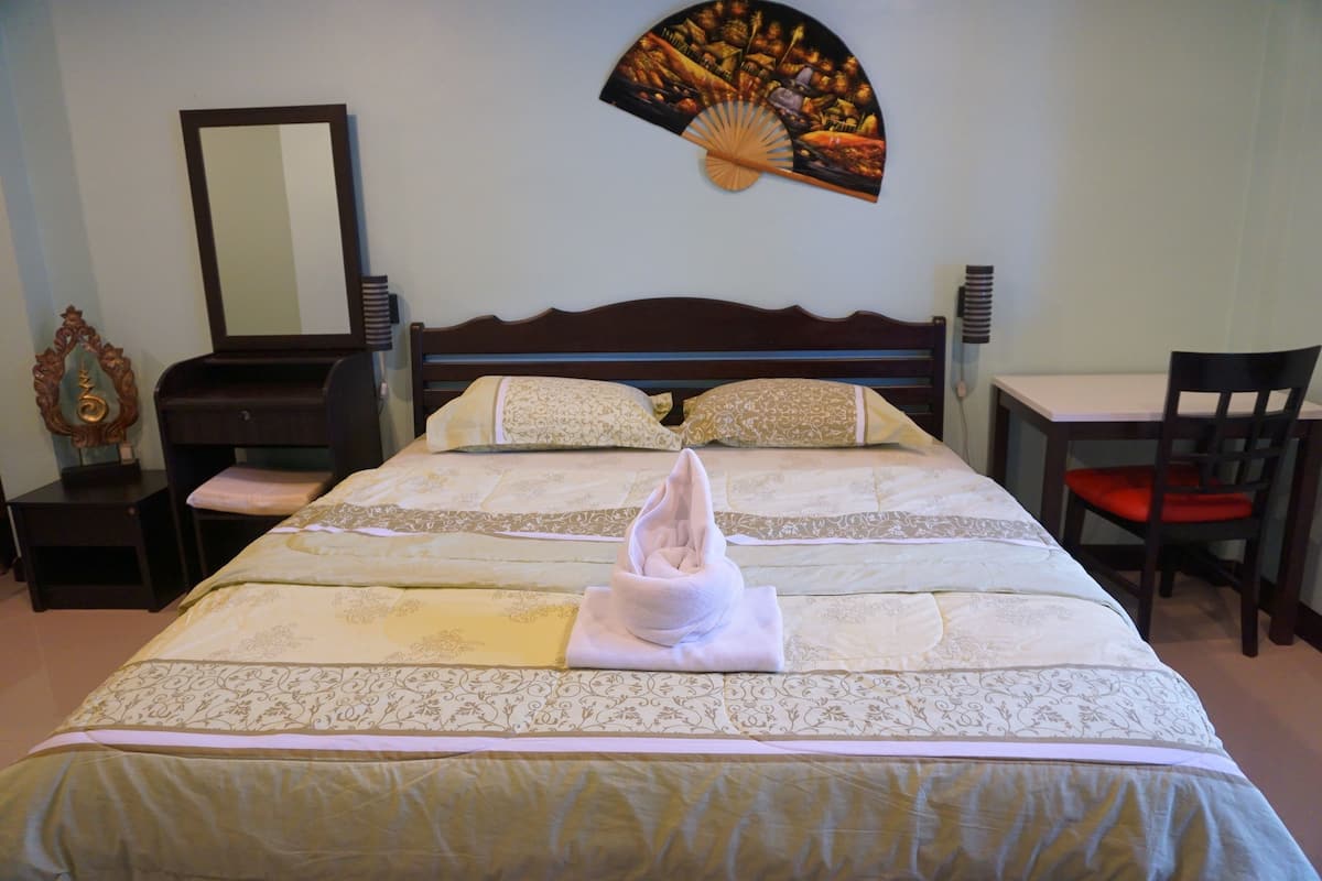 Een opgemaakt bed met een decoratieve handdoek, een houten hoofdbord, een ventilator aan de muur, een bureau en een stoel.