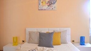 Un dormitorio con cabecera blanca, una cama con muchos almohadones, una mesita de noche con lámpara y un cuadro en la pared.