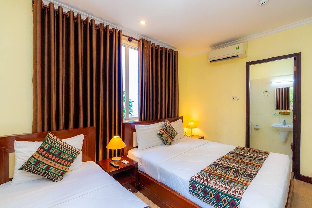 Une chambre d’hôtel avec deux lits, une table de chevet avec une lampe, une fenêtre avec des rideaux, un appareil de climatisation et une salle de bain avec un lavabo et un miroir.