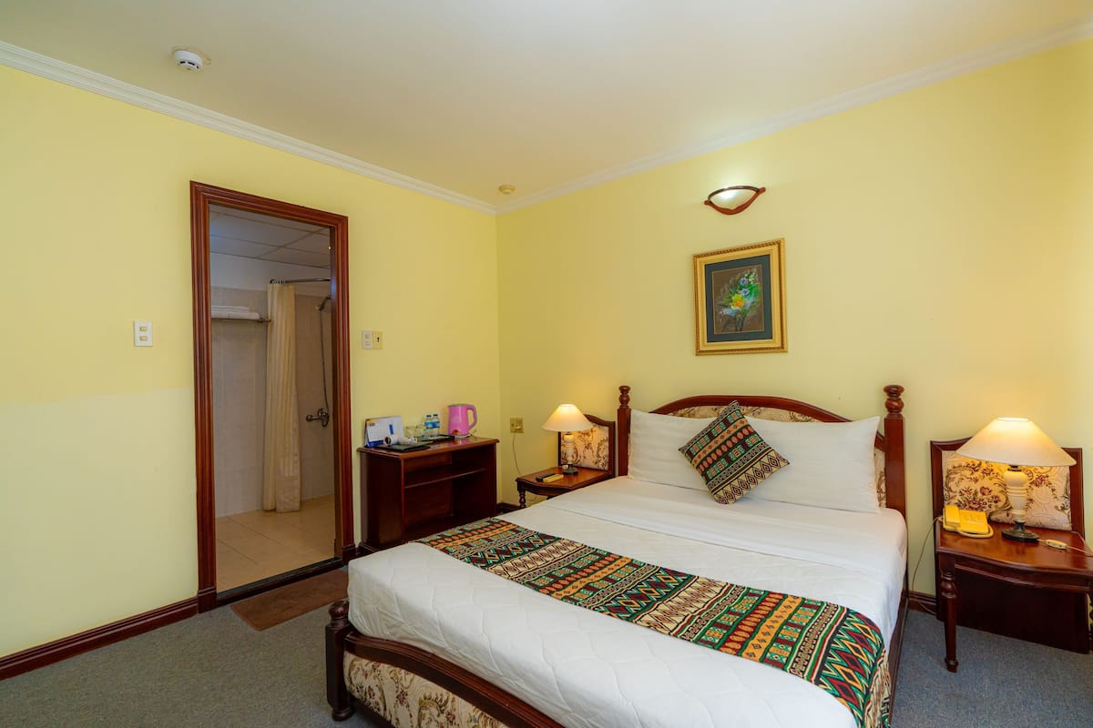 A hotel room with a large bed, two bedside tables, a nightstand, a mirror, a painting, and a door leading to a bathroom.
