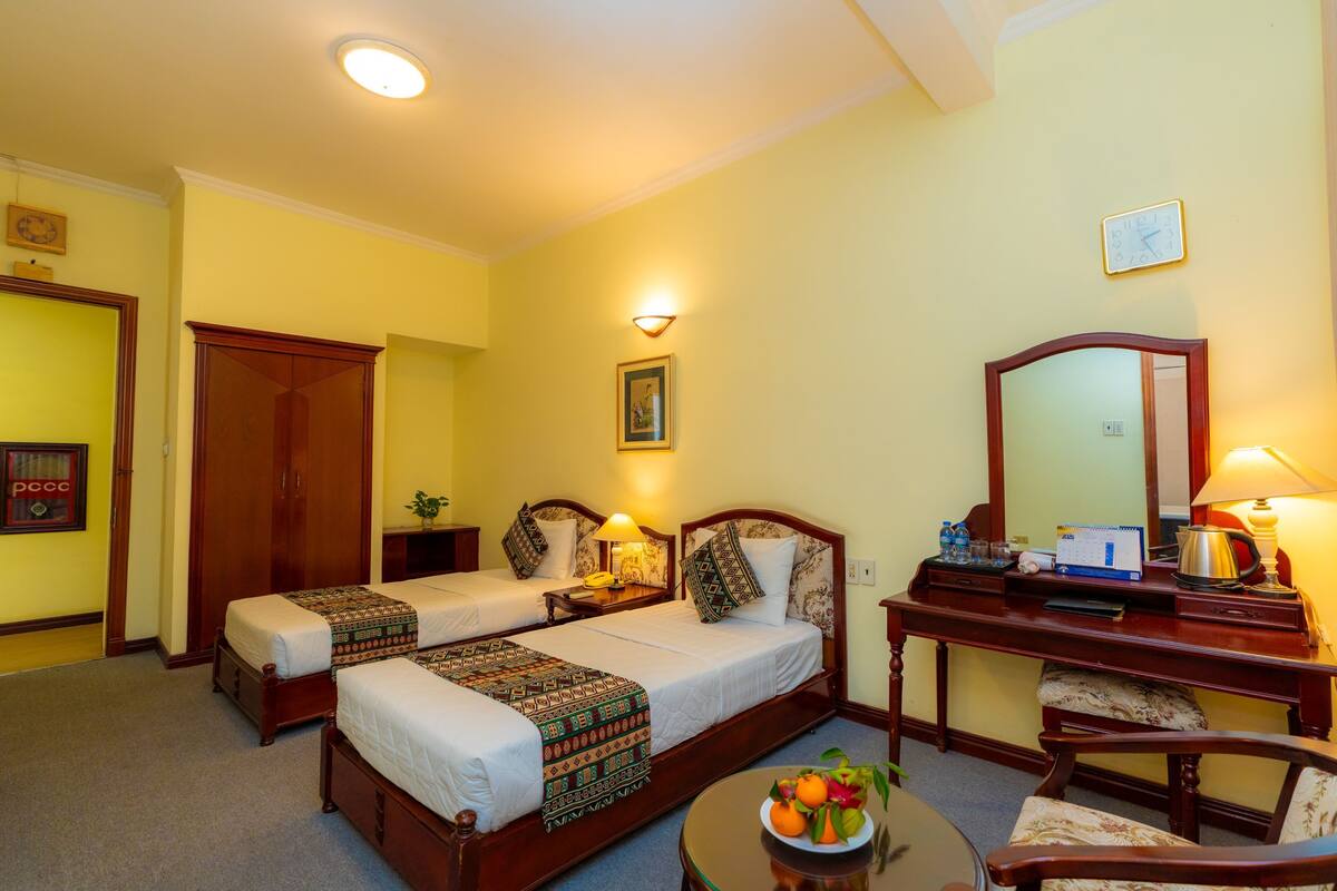 Une chambre d’hôtel avec deux lits simples, un bureau avec une lampe, une chaise, une table ronde avec des fruits et un miroir.