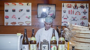 Un chef vêtu d’une blouse blanche et d’une casquette à motifs, se tient derrière un comptoir où sont disposées des bouteilles de saké et un service à thé japonais traditionnel.