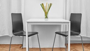 Um quarto com uma mesa branca, duas cadeiras pretas, um relógio amarelo na parede e um vaso com lápis sobre a mesa.