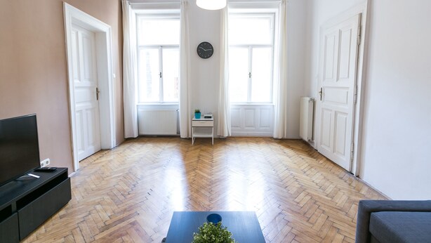 Un salon spacieux doté d’un parquet en bois à motifs en chevrons, de portes blanches, d’une horloge noire et d’un luminaire suspendu.