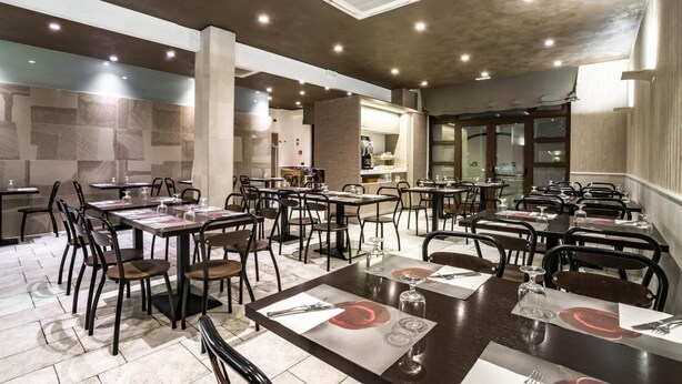 A restaurant with multiple tables and chairs, a kitchen area in the background, and a modern ceiling design. - Hotel New Bari (Bitritto)