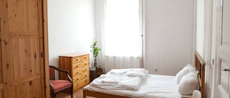 Un dormitorio con una cama de madera, un armario de madera, una silla, una mesita de noche con una lámpara y una ventana con cortinas translúcidas.