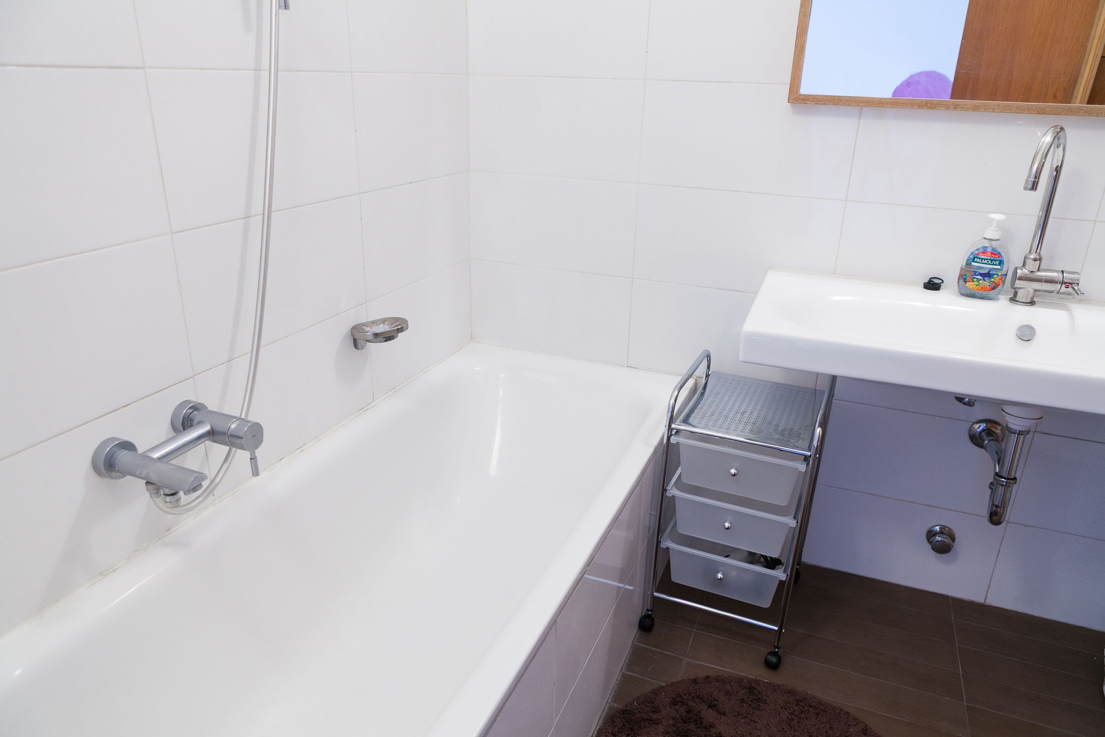 A bathroom with a white bathtub, a sink with a soap dispenser, and a metal rack with drawers.