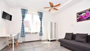 Una sala de estar moderna con un sofá gris, un escritorio blanco con una silla, un ventilador de techo y un televisor montado en la pared.