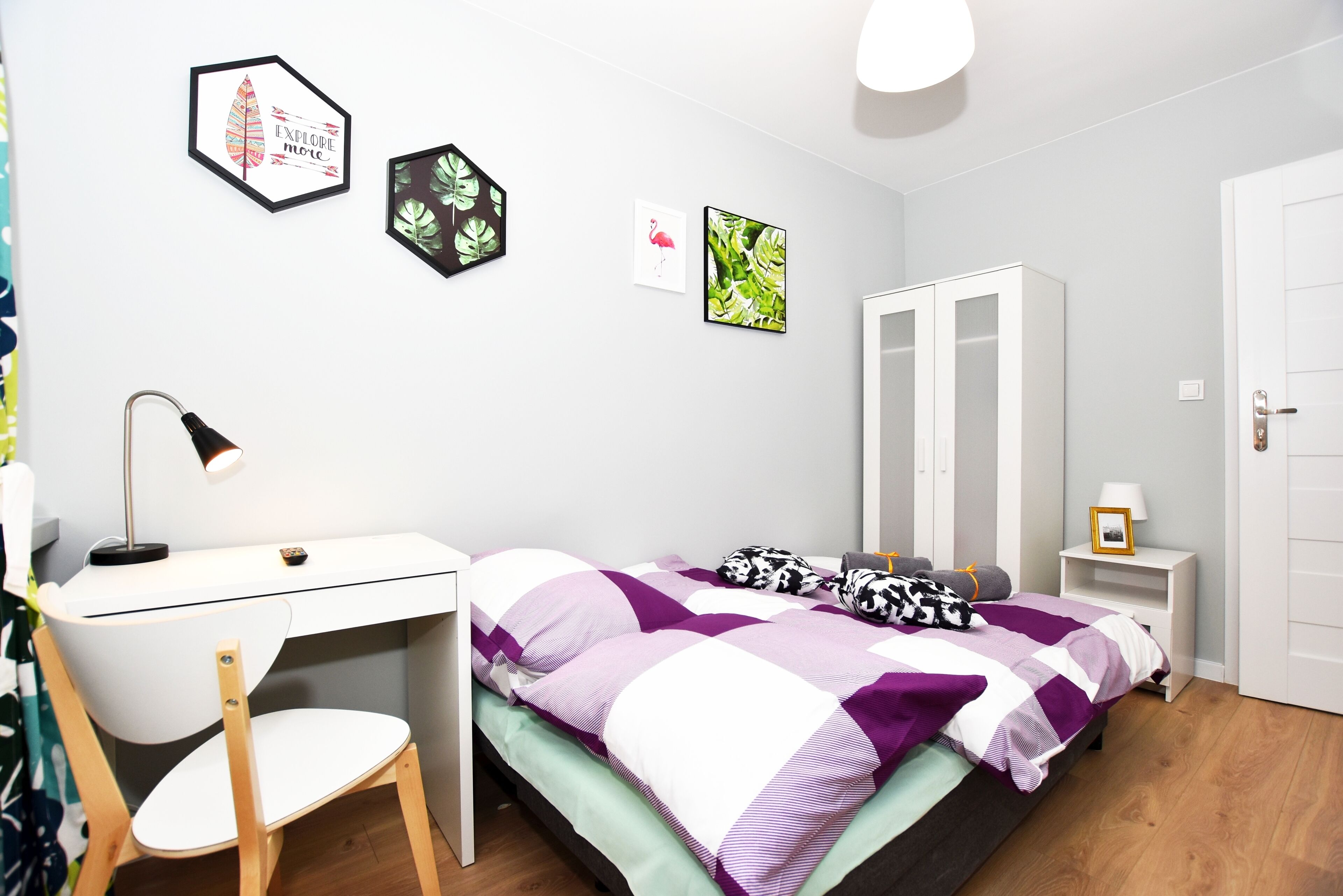 A bedroom with a bed, desk, chair, wardrobe, and wall art.