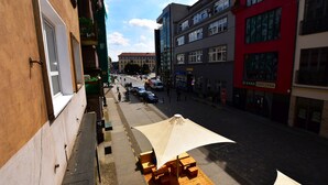 Vista de la calle con una fila de mesas al aire libre bajo un toldo, un edificio con un cartel que dice "KASA STEFCIYKA", y un paseo peatonal.