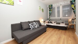 Una sala moderna con un sofá gris, una cama pequeña, piso de madera y una ventana con cortinas traslúcidas.