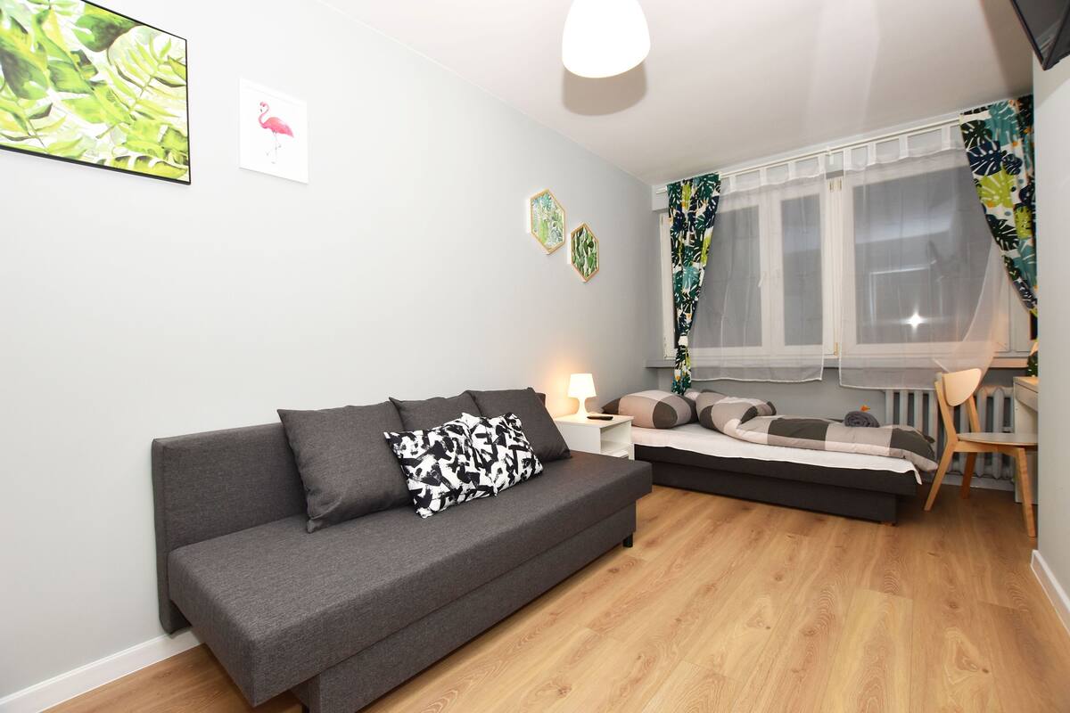 A modern living room with a grey sofa, a small bed, a wooden floor, and a window with sheer curtains.