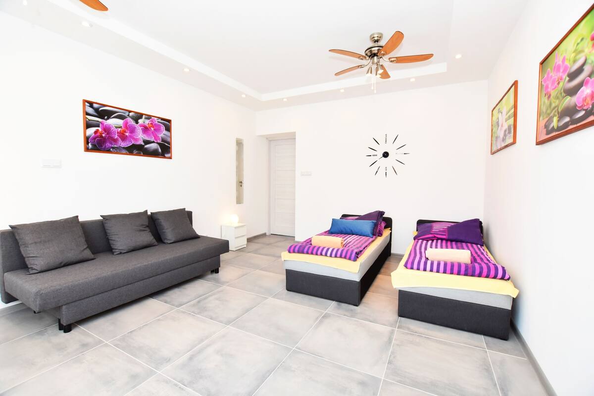 A modern room with a grey sofa, two beds with colorful bedding, a ceiling fan, and artwork on the walls.